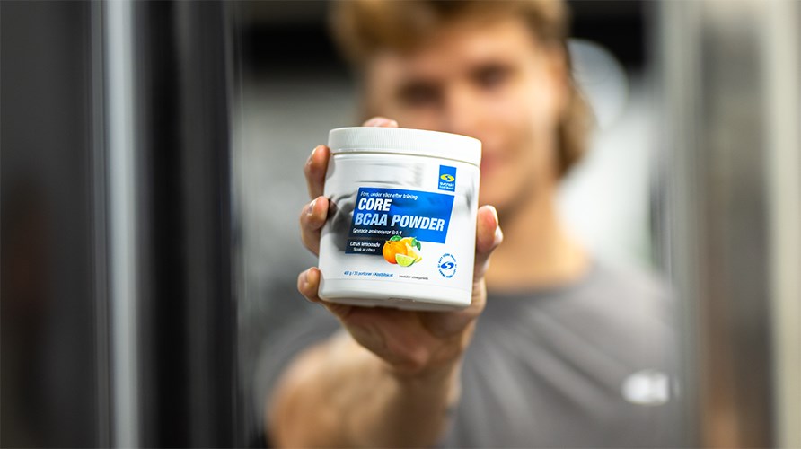 Henkil pitelee Core BCAA Powder -purkkia kameraa kohti. Henkil ja tausta ovat eptarkkoja.