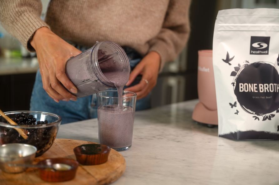 Nainen kaataa mustikkasmootieta lasiin. Taustalla Healthwell Bone Broth -luuliemijauhepussi ja etualalla kulhollinen mustikoita.