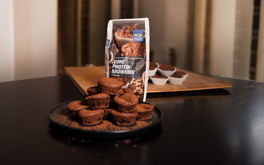 Valmiiksi paistettuja proteiinibrownieita lautasella, taustalla pussi Core Protein Brownies -leivontaseosta.