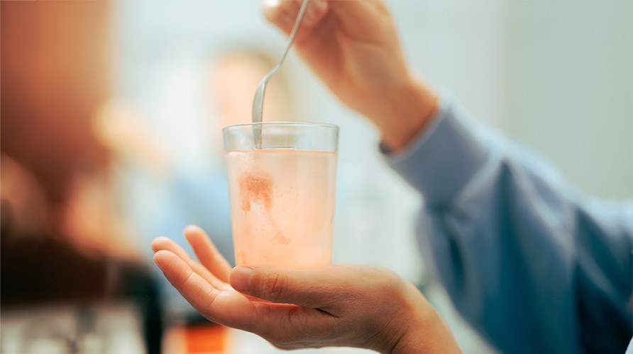 K�si pit�� vesilasia alhaalta ja toinen k�si sekoittaa elektrolyyttijauhetta vedess� lusikan avulla. Tausta on sumea.