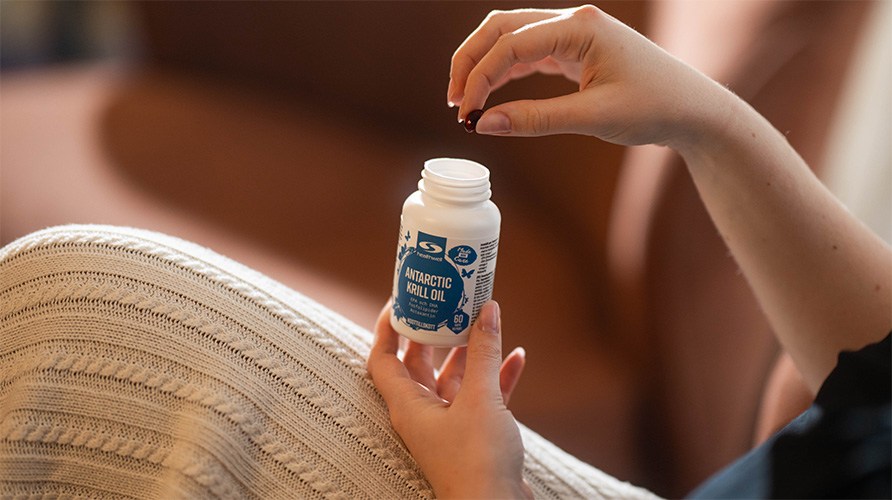 Nainen, p��ll��n vaalea neulehame, pitelee Healthwell Antarctic Krilli�ljy -purkkia nosten sielt� yhden kapselin.