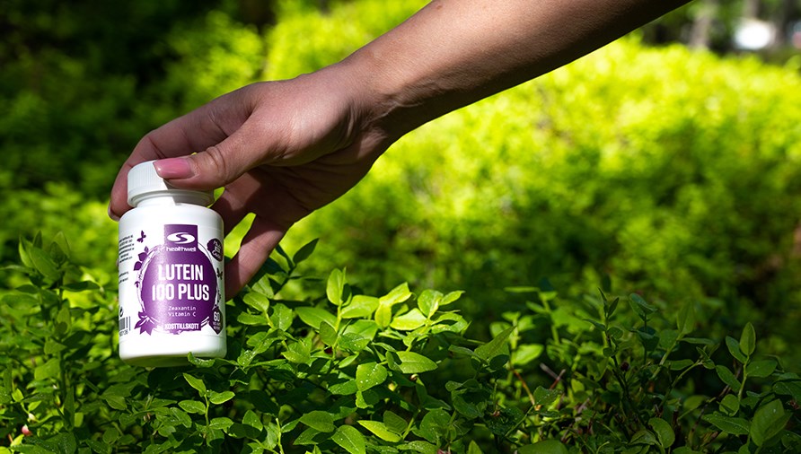 Ksi pit Healthwellin Lutein Plus -purkkia, taustalla vihre pensas.