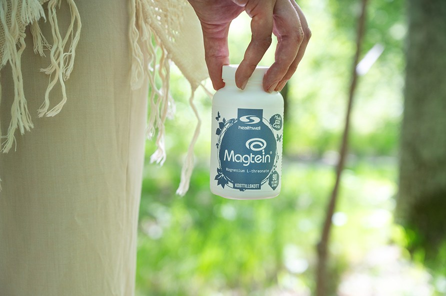 Naisen sormet roikottavat Healthwell Magnesium Magtein -purkkia metsist taustaa vasten.