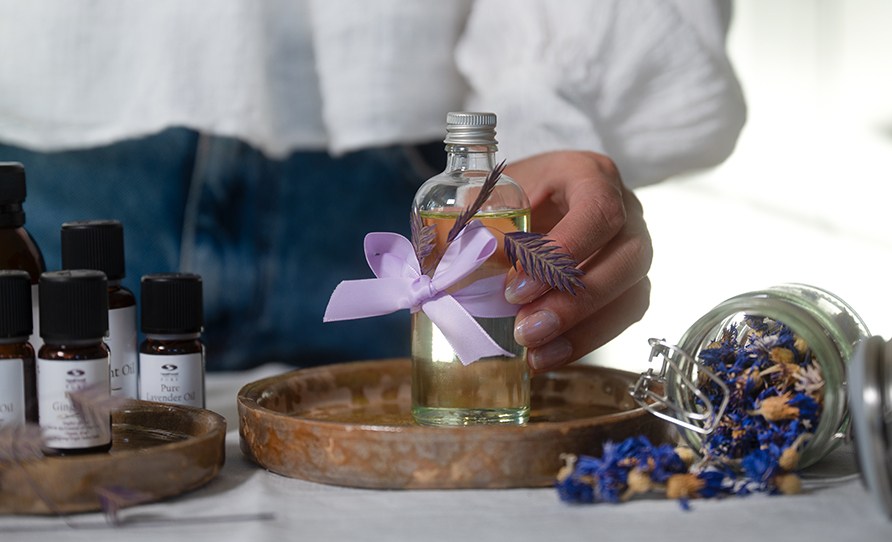 Nainen ottamassa lasista hierontaöljypulloa keraamiselta tarjottimelta. Pöydällä myös eteerisiä öljyjä ja kuivattuja kukkia purkissa. Nainen ottamassa lasista hierontaöljypulloa keraamiselta tarjottimelta. Pöydällä myös eteerisiä öljyjä ja kuivattuja kukkia purkissa.