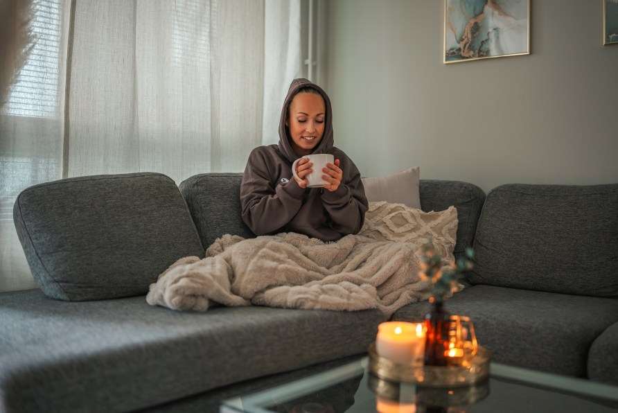 Nainen istuu sohvannurkassa viltin alla ksissn teemuki.