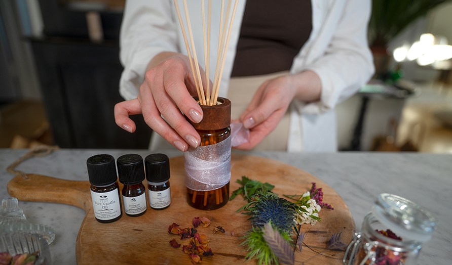 Nainen ottamassa lasista puutikuilla varustettua tuoksupulloa puiselta leikkuulaudalta, jolla my�s eteerisi� �ljyj� ja kuivakukkia.